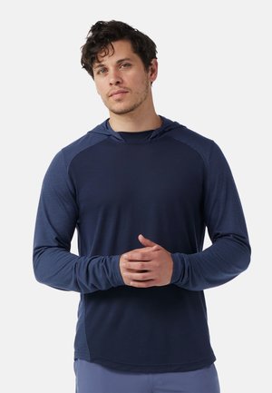 Man met kort krullend haar, gekleed in een marineblauwe hoodie met patroonmouwen, staand met gevouwen handen voor zich tegen een witte achtergrond.