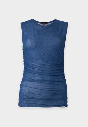 Femme aux cheveux blond clair portant un haut sans manches bleu marine, un jean large bleu clair et des bottes noires à talons, debout devant un fond blanc uni.