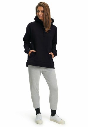 Sweatshirt à capuche noir avec poches avant, associé à un pantalon de survêtement gris cintré. Modèle portant des baskets blanches. Design simple et fonctionnel.