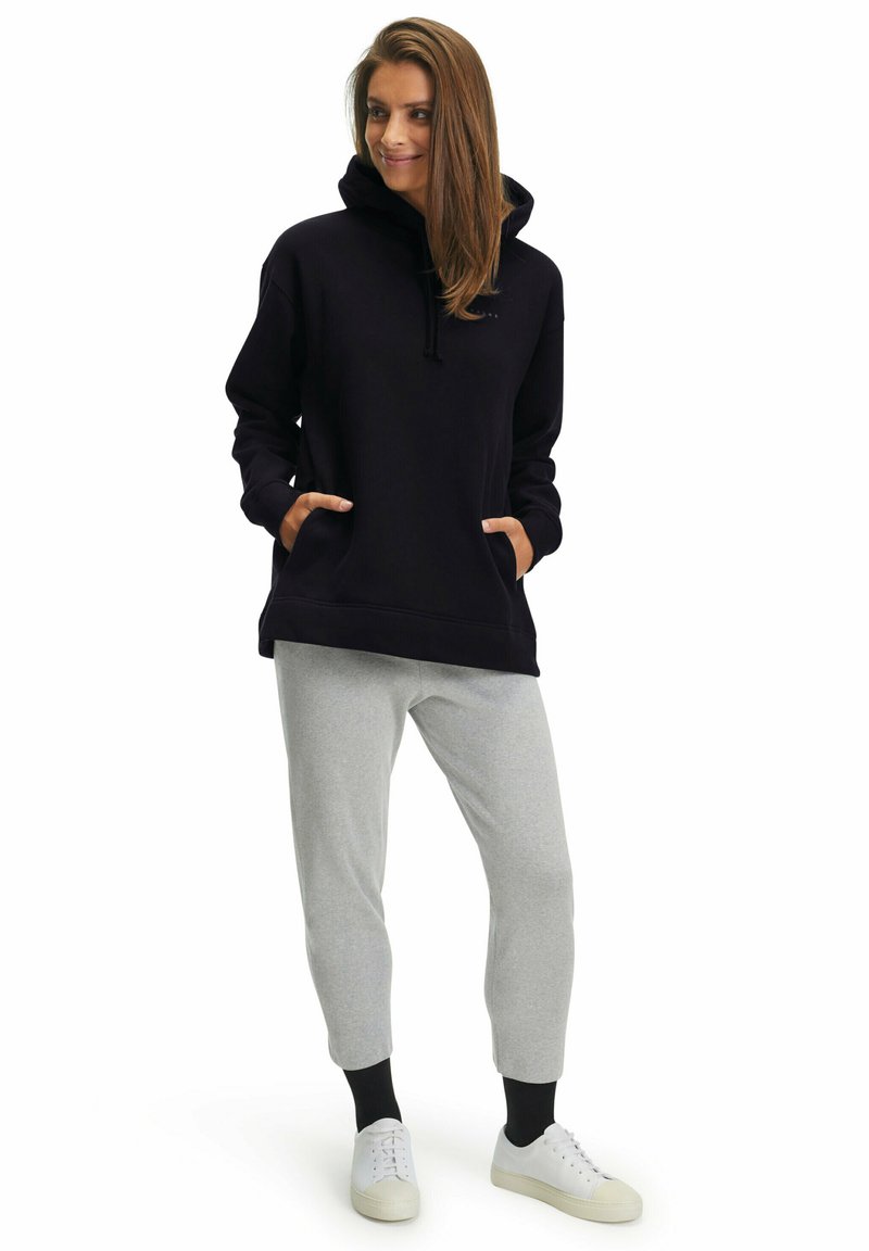 Felpa nera con cappuccio e tasche frontali, abbinata a pantaloni sportivi grigi a gamba affusolata. Modello che indossa scarpe bianche. Design semplice e funzionale.