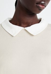 Beige gebreide trui met een witte, ronde kraag. De stof is gestructureerd met een fijn patroon, wat de details van de halslijn en kraag benadrukt.