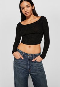 Fekete, hosszú ujjú crop top csillogó anyagból, magas derekú kék farmernadrággal párosítva. A felső kerek nyakkivágással és szűkített ujjal rendelkezik.