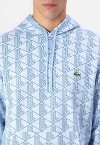 Detailní záběr na osobu v světle modré mikině s kapucí, která má geometrické vzory a malé zelené logo krokodýla na hrudi.