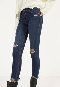 Mujer vistiendo pantalones ajustados rasgados de color azul oscuro con dobladillos deshilachados, combinados con botas negras hasta el tobillo y un suéter blanco desgastado.