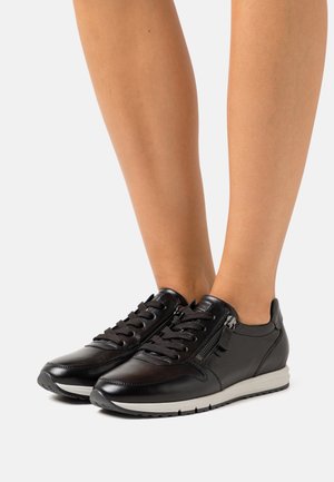 Black leather lace-up sneakers with side zippers worn on bare legs, featuring white and black rubber soles on a light background.