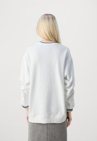 Sudadera blanca de gran tamaño con mangas largas, que presenta un cuello y puños acanalados con una franja oscura. Textura suave, corte holgado.