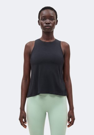 Kvinde stående med fronten mod kameraet iført en ærmeløs sort top og lysegrønne leggings mod en ensfarvet baggrund.