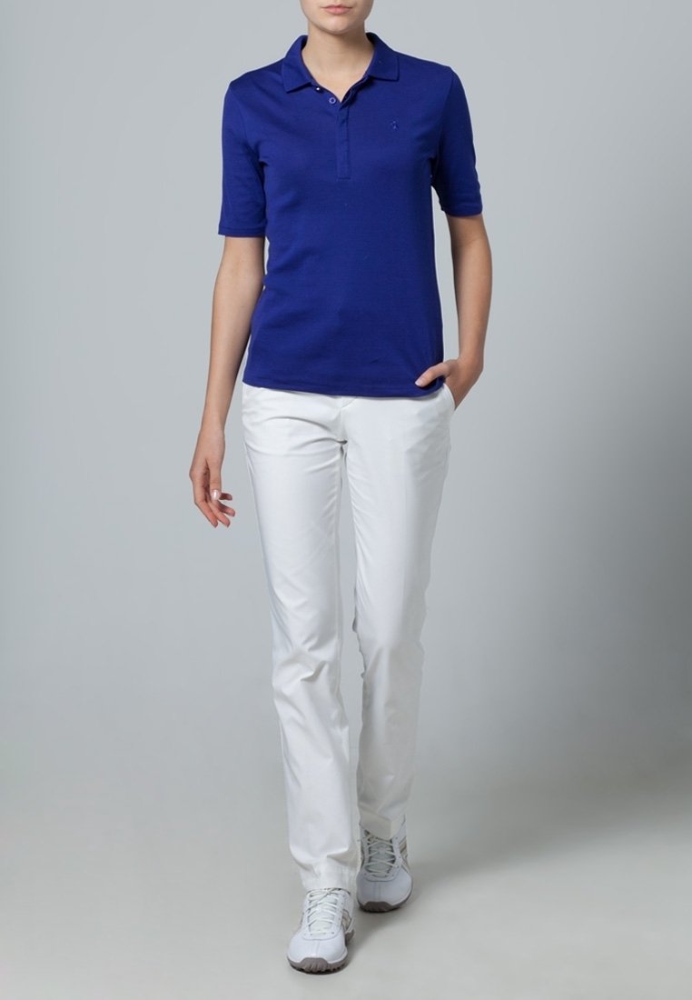 Personne debout avec une main dans la poche, portant un polo bleu, un pantalon blanc et des baskets blanches sur un fond gris uni.