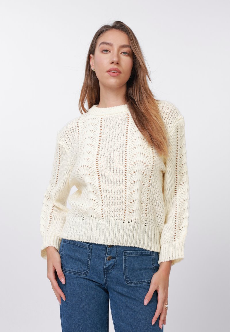 Lyst cremefarvet strikket sweater med åben maskestrik, rundet halsudskæring og ribbet kant. Kombineret med blå denimjeans.