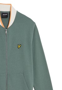 Veste zippée vert clair avec un col contrastant blanc et orange, dotée d'un logo d'oiseau jaune brodé sur la poitrine et de poches avant.