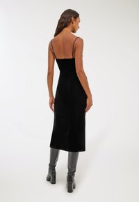 Czarna aksamitna sukienka midi z cienkimi ramiączkami, dopasowaną górą i gładką fakturą, zestawiona z czarnymi kozakami na wysokim obcasie sięgającymi do kolan.