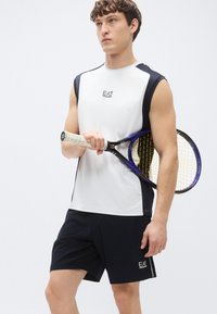 Biele športové tielko bez rukávov s tmavomodrými bočnými panelmi a logom, kombinované s tmavomodrými šortkami. Model drží tenisovú raketu s textúrovanou rukoväťou.