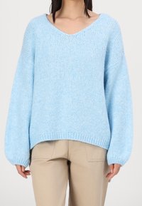 Femme portant un pull en tricot léger bleu clair, ample, avec de longues manches ballon, et un pantalon beige, debout devant un fond uni.