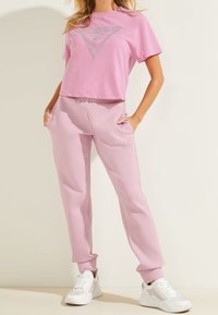 Person, die ein pinkfarbenes Cropped-T-Shirt und passende pinkfarbene Jogginghosen mit weißen Sneakers trägt, steht mit den Händen in den Taschen.