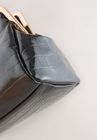 Melna rokassoma ar krokodila ādas reljefu un spīdīgu virsmu, rotāta ar zelta detaļām un strukturētu formu. Redzama teksturēta virsmas detaļa.