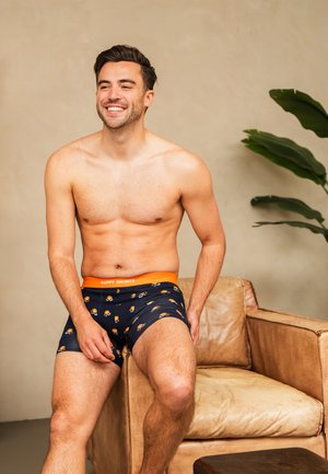 Lachende man zonder shirt zittend op een beige leren stoel, gekleed in marineblauwe boxershorts met oranje cartoonprints en een oranje tailleband.