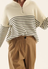 Pull en côte crème avec des rayures horizontales noires, doté d'un col à fermeture éclair quart. Associé à un pantalon plissé marron taille haute.