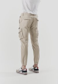 Pantalones cargo beige con piernas estrechas, múltiples bolsillos y puños elásticos, combinados con zapatillas claras que presentan detalles en rojo, negro y gris.
