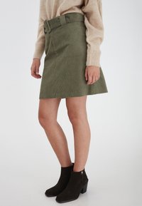 Olivgrön kordfläskkjol med ett brett bälte, A-linjeform, ribbad textur och rå nederkant, som matchas med mörka mocka ankelskor.