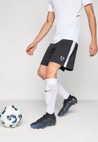 Fekete focirövidnadrág fehér oldalsó díszítéssel, fehér atlétatrikóval, fekete-kék stoplis cipővel és fehér zoknival párosítva. Egy focilabda van a közelben.