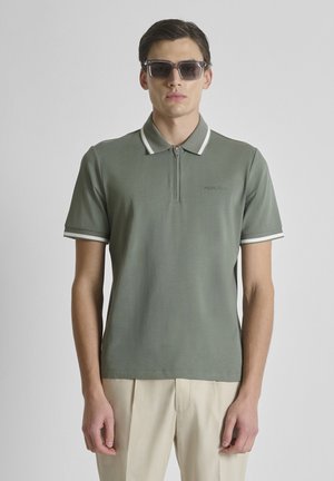 Hombre con camisa polo de manga corta verde con ribete blanco y pantalones beige, mirando hacia adelante con gafas de sol rectangulares oscuras.