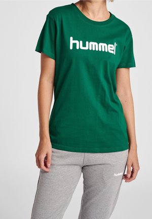 Personne portant un t-shirt vert Hummel et un pantalon de survêtement gris Hummel, debout, les bras détendus le long du corps.