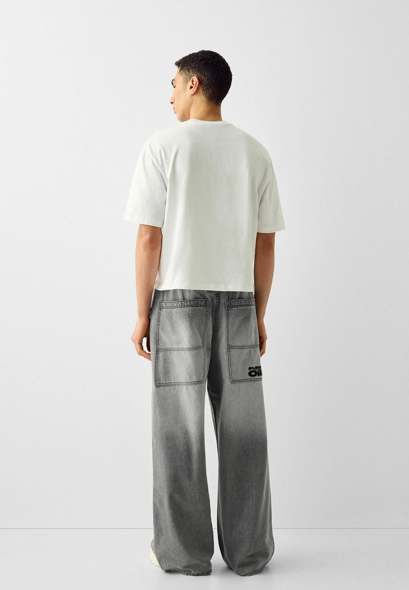 Uomo con una t-shirt bianca oversize e jeans grigi larghi con ampie tasche posteriori e dettagli in testo nero, in piedi su uno sfondo bianco uniforme.