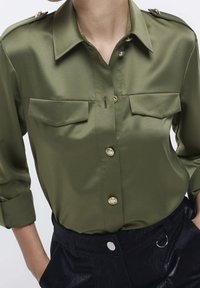 Olivgrön satin skjorta med knappar, krage, två bröstfickor och guldtonade knappar. Har uppvikta ärmar och en mjuk yta.