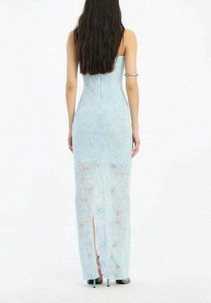 Mujer con cabello largo y oscuro que lleva un vestido de encaje azul claro sin tirantes con una abertura en la parte trasera y tacones altos azules, de espaldas.
