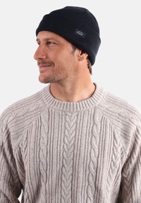 Bonnet tricoté bleu marine avec une texture côtelée et une petite étiquette de marque. Porté avec un pull en tricot torsadé beige clair.