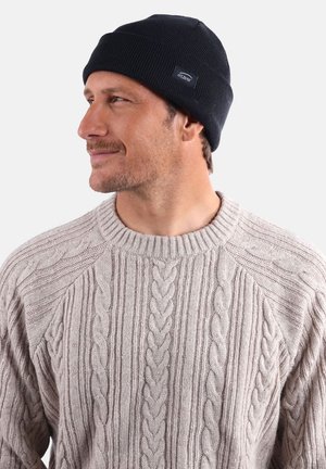 Bonnet tricoté bleu marine avec une texture côtelée et une petite étiquette de marque. Porté avec un pull en tricot torsadé beige clair.