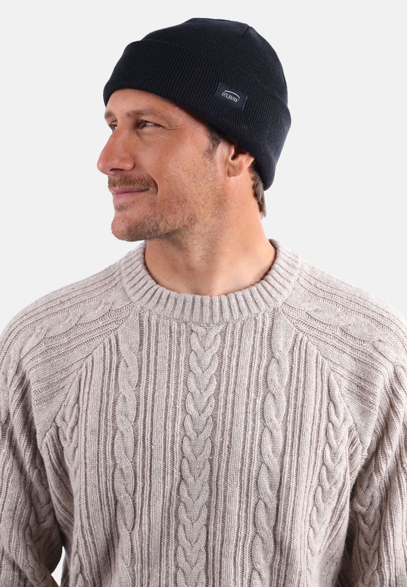 Bonnet tricoté bleu marine avec une texture côtelée et une petite étiquette de marque. Porté avec un pull en tricot torsadé beige clair.