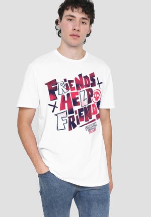 Young man wearing white T-shirt with red and blue text "Friends Help Friends" and small skull graphic, paired with blue jeans.