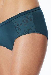 Culotte teal a vita media realizzata in un tessuto liscio con un motivo floreale in pizzo sui lati. Presenta un'ampia fascia in vita per comfort e supporto.