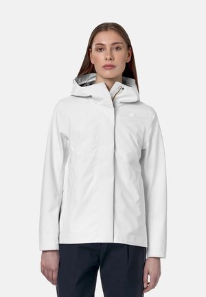 Kvinde iført en hvid hættejakke med lynlås foran og lange ærmer, kombineret med mørke bukser, stående mod en ensfarvet baggrund.