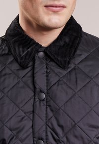 Casaco preto acolchoado com colarinho de veludo cotelê, botões de pressão e uma textura suave, destacando o padrão de losangos pelo tecido.