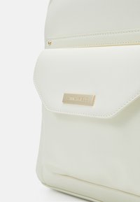 Sac à dos blanc en matériau synthétique lisse, avec une poche à rabat, des accessoires en métal doré et une étiquette de marque rectangulaire embossée à l'avant.