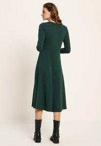 Frau mit roten Haaren, die ein langärmliges, dunkelgrünes Midikleid und schwarze Knöchelstiefel trägt, steht vor einem schlichten beigen Hintergrund.