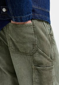 Olivengrønne kordfløyelbukser med vertikale riller, med sidelommer og en lapp-lomme på baksiden, supplert med en denimjakke.