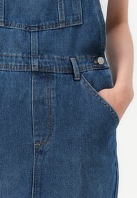 Sinine teksariidest üleüldine, millel on eesmine tasku, dekoratiivne õmblus ja metallist nööbi aktsent. Kangas näeb välja tugev ja tekstureeritud.