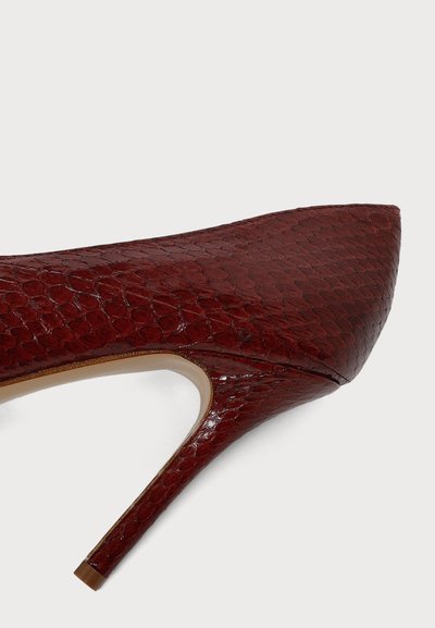 Chaussure à talon haut rouge foncé avec bout pointu et motif peau de serpent texturé, vue de côté sur un fond clair uni.