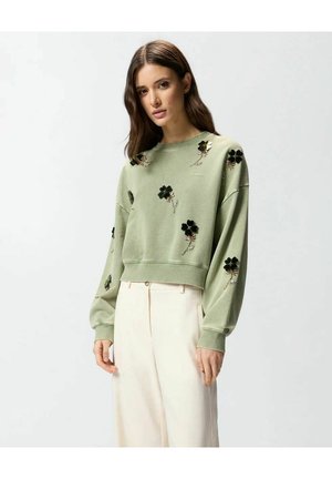 Donna che indossa una felpa verde chiaro con ricami floreali neri abbinata a pantaloni a gamba larga color crema e a vita alta, in piedi davanti a uno sfondo semplice.