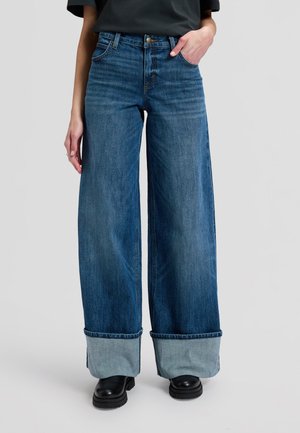 Person trägt blaue Jeans mit weitem Bein und großen umgeschlagenen Säumen sowie schwarze Stiefel, steht vor einem schlichten Hintergrund.