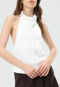 Witte mouwloze haltertop met een coltrui-halslijn en een gladde textuur. Heeft open zijkanten en een losvallende pasvorm, gecombineerd met een donkere stof eronder.