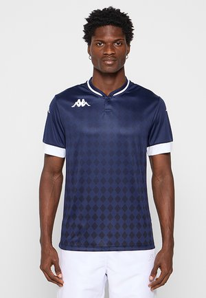 Uomo in piedi di fronte indossa una maglietta sportiva blu navy a motivo a diamante con rifiniture bianche sulle maniche e pantaloncini bianchi.