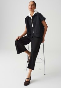 Black cropped vest with buttons, paired with black wide-leg pants. A white hoodie is visible underneath. The shoes are black mesh with a strap.