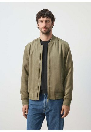 Mann mit kurzen lockigen Haaren, der eine olivgrüne Bomberjacke, ein schwarzes Shirt und blaue Jeans vor einem schlichten hellen Hintergrund trägt.