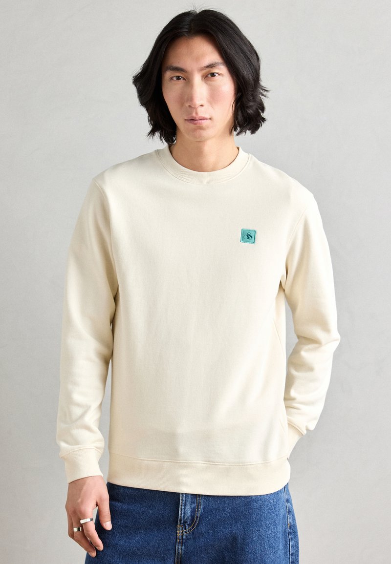 Crème kleur sweatshirt van zacht materiaal met een ronde hals. Beschikt over een klein turquoise logo op de borst en ribgebreide manchetten en zoom.