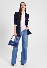 Blazer blu navy, camicia bianca con cravatta e jeans a gamba larga. Borsa in pelle blu con motivo coccodrillo e dettagli dorati. Scarpe con tacco alto e accenti dorati.