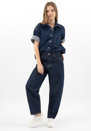 Giovane donna che indossa una camicia di jeans blu scuro con maniche arrotolate e jeans a gamba larga abbinati, posando con le mani nelle tasche e sneakers bianche.